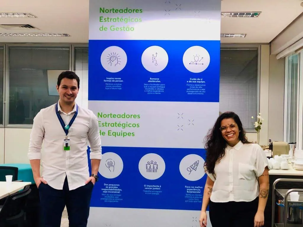 Bruna e um colega em frente a um painel azul com os norteadores estratégicos de gestão e de equipes do Agibank