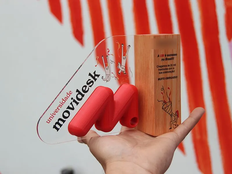 Troféu de acrílico e madeira com o logo da Universidade Movidesk, segurado na palma da mão