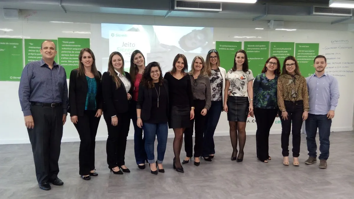 Grupo de treze pessoas em uma sala de treinamento do Sicredi, com cartazes verdes ao fundo