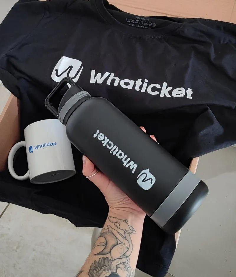 Mão segurando uma garrafa térmica preta com o logo da Whaticket, sobre uma camiseta preta e ao lado de uma caneca branca da marca